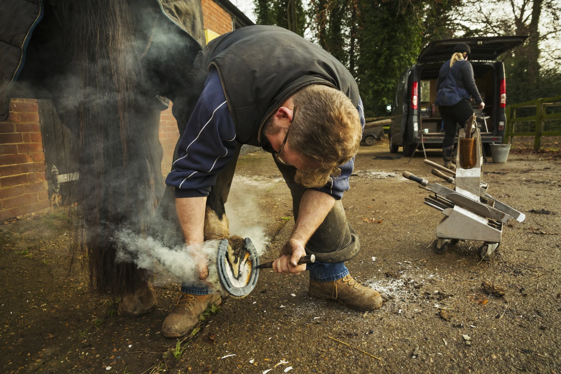 A farrier shoeing a horse, bending down and fitting a new horseshoe to a horse's hoof.